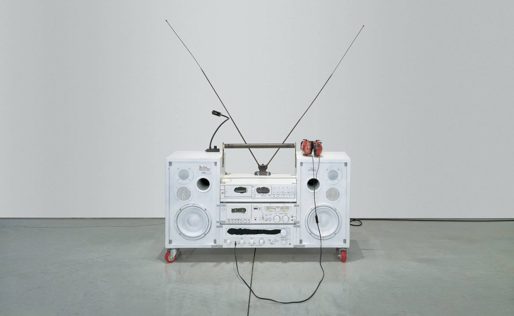 Sculpture depicting a boombox painted mostly white on small red casters in a neutral gallery space.